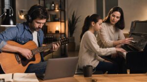Self taught guitarist practicing near a laptop while a teacher guides a piano student